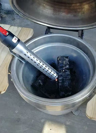 A stainless steel charcoal grill with its lid open, showing black charcoal coals inside. A handheld electric lighter with a silver body and black handle is being used to ignite the coals.