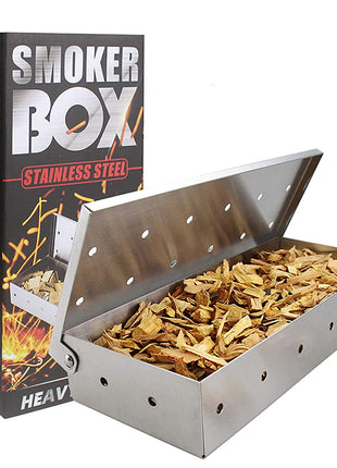 A stainless steel smoker box filled with wood chips, shown next to its black retail box labeled 'SMOKER BOX STAINLESS STEEL'.